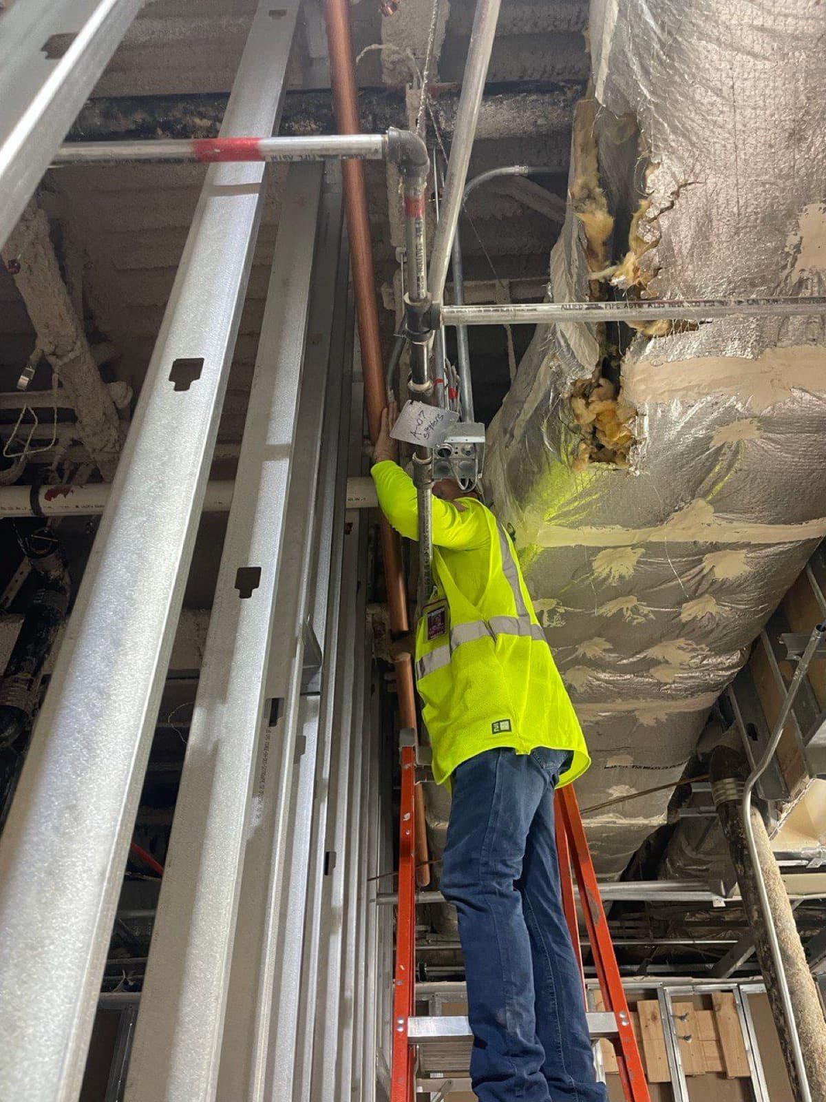 Plomero de RCR en escalera instalando tubería colgada del techo